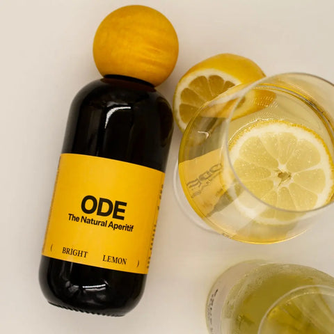 ODE Bright Lemon Aperitif liegend mit zwei gefüllten ODE Lemon Spritz Gläsern und halber Zitrone, Sommerdrink